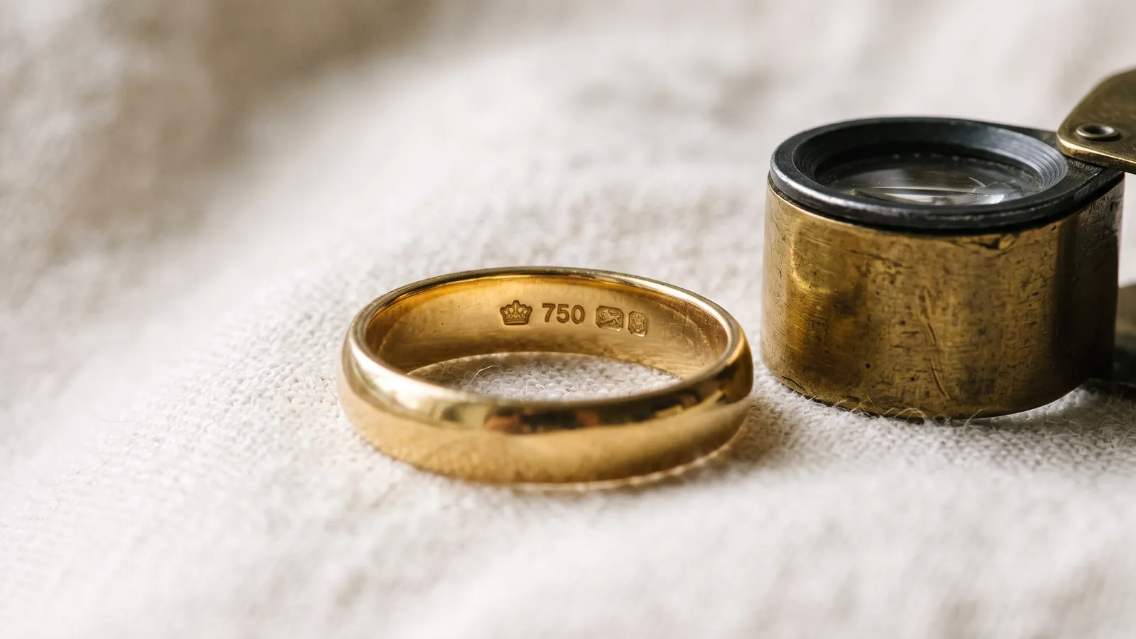 Ehering aus Gelbgold auf cremefarbenem Leinen neben einer Juwelierlupe